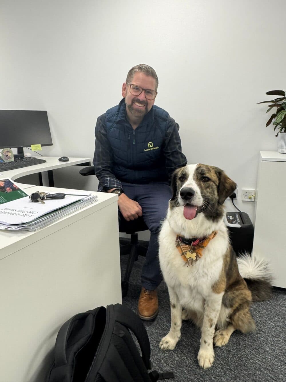 Man sitting at a desk in an office, smiling, with a large dog sitting beside him on the floor. - Home Instead
