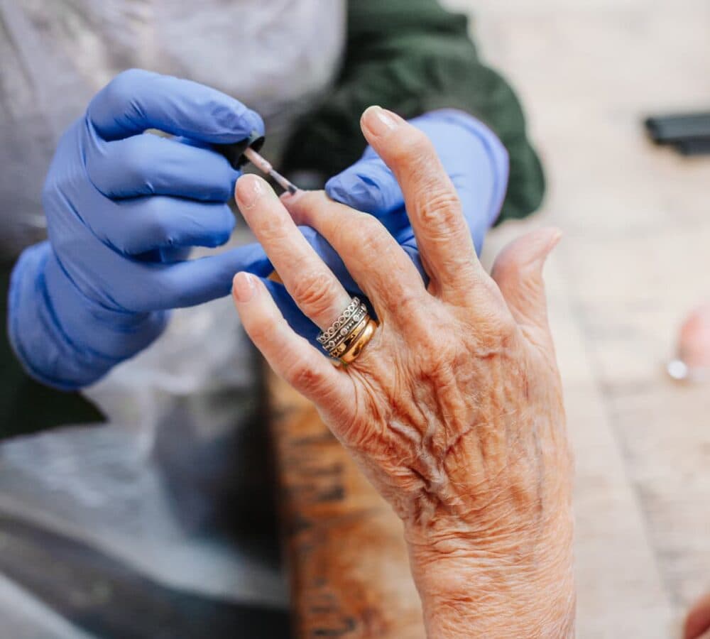An old woman wearing rings and having her nails done with a carer wearing blue gloves - Home Instead