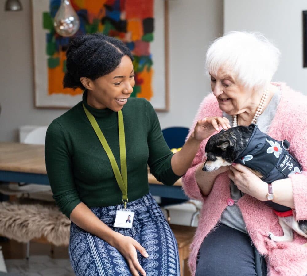 An old female adult with white hair and wearing pink happy and smiling while holding her dog and with her female carer with black hair and wearing green inside the house