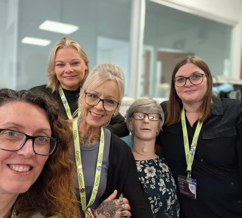 Four women smile with a medical training mannequin, all wearing lanyards, in a bright classroom. - Home Instead
