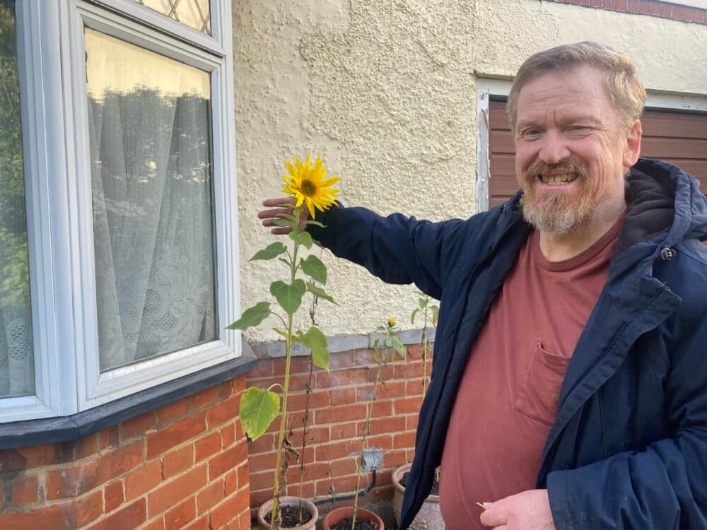 A smiling man stands beside a sunflower growing in a pot outside a house, gently touching the flower. - Home Instead