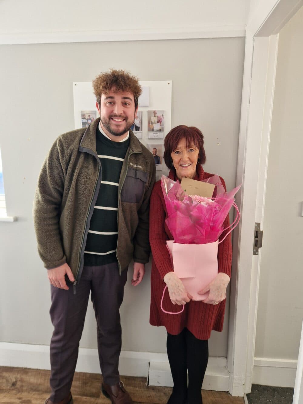 A smiling man stands next to a woman holding a bouquet of pink flowers in a gift bag, indoors. - Home Instead