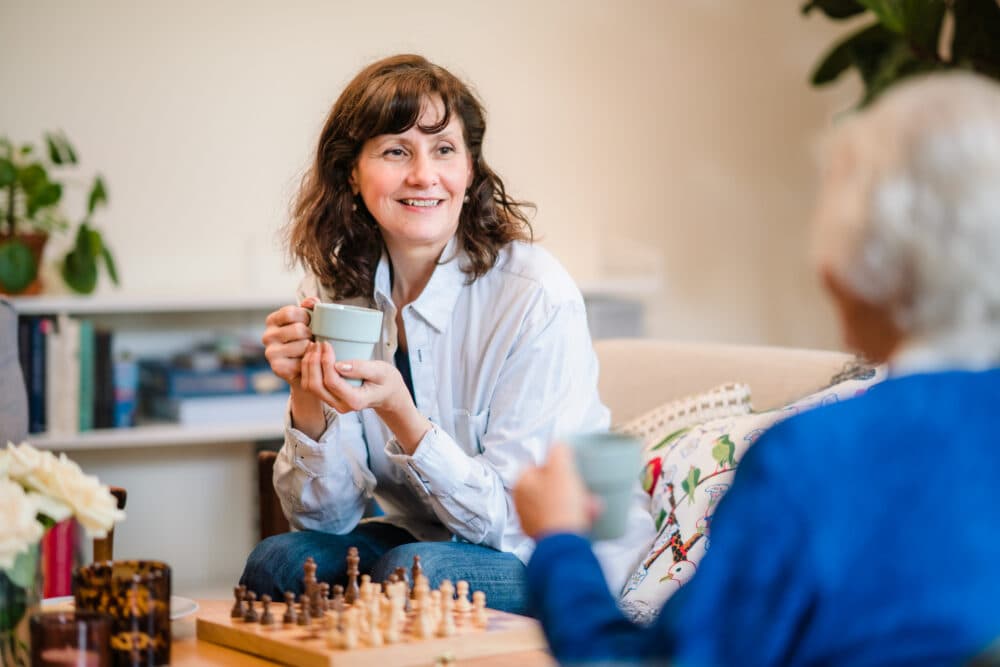 A woman smiles, holding a mug, whilst talking to another person near a chessboard in a cosy room. - Home Instead