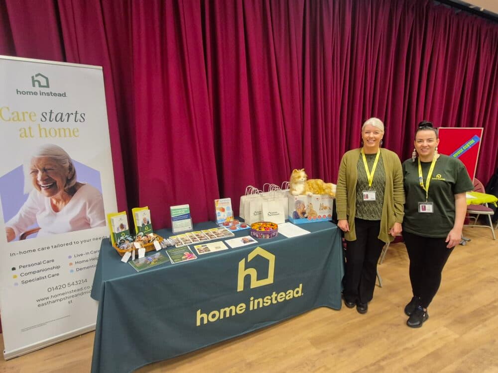 Two women stand by a "Home Instead" care service booth with leaflets and a cat plush, in front of a red curtain. - Home Instead