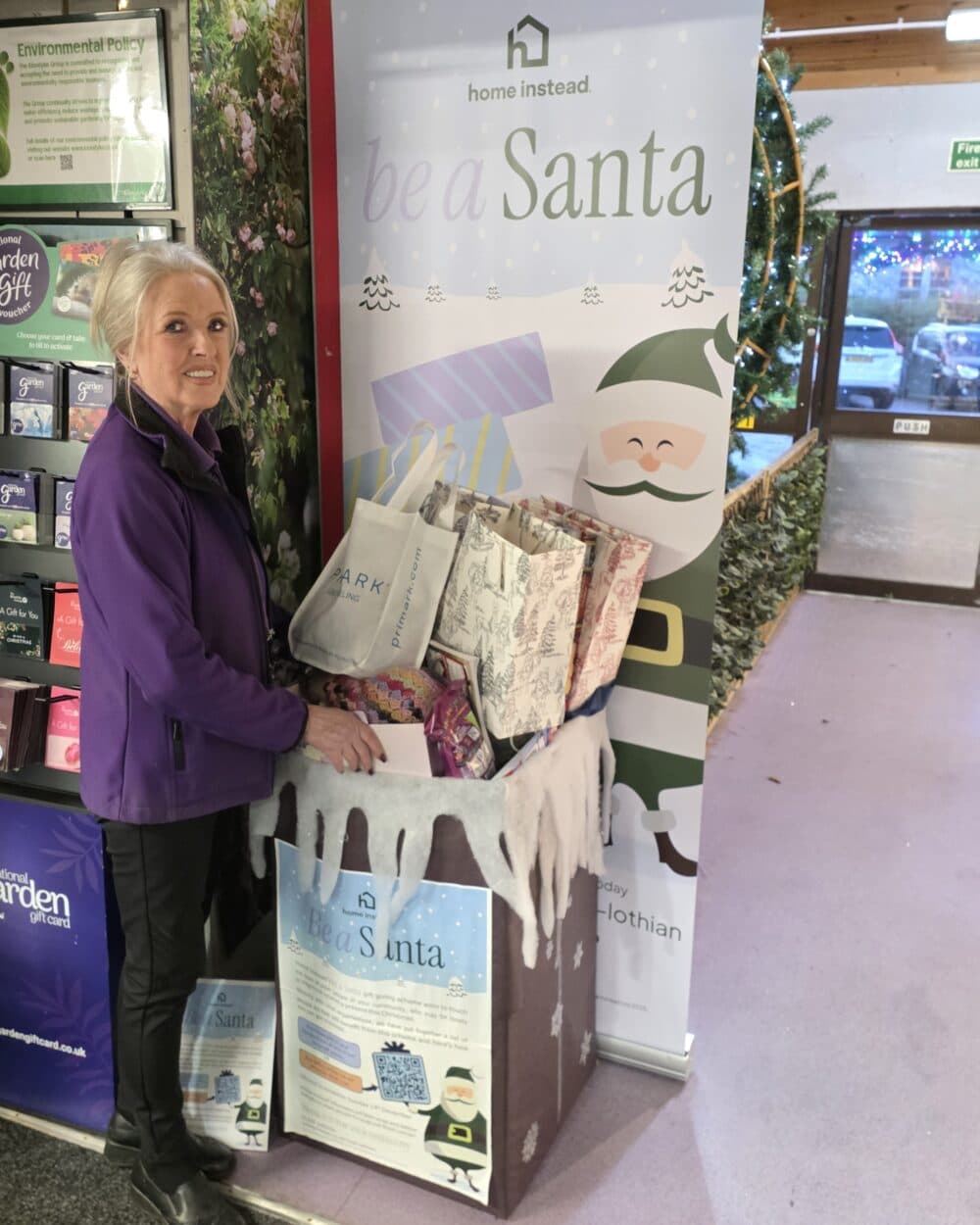 A woman standing by a festive "Be a Santa" donation box filled with gift bags and presents. - Home Instead