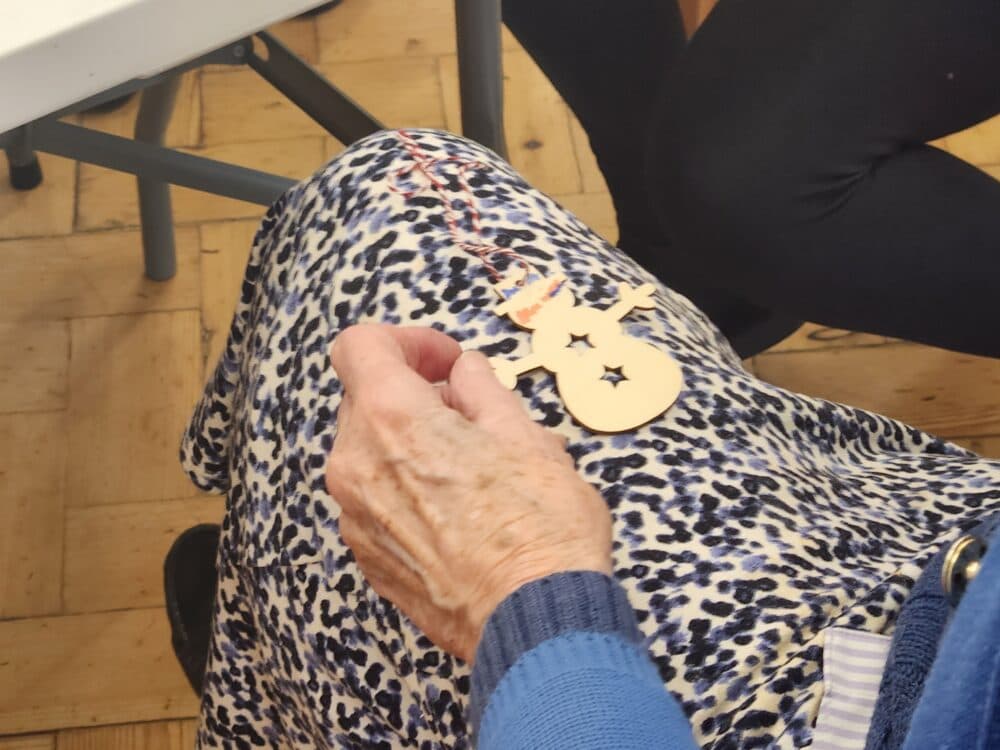 An elderly hand holds a wooden snowman ornament on a lap covered by a blue patterned skirt. - Home Instead