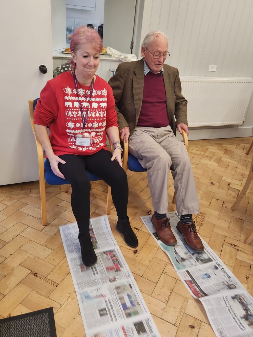 A woman and an older man sit on chairs with their feet on newspaper sheets spread on the floor. - Home Instead