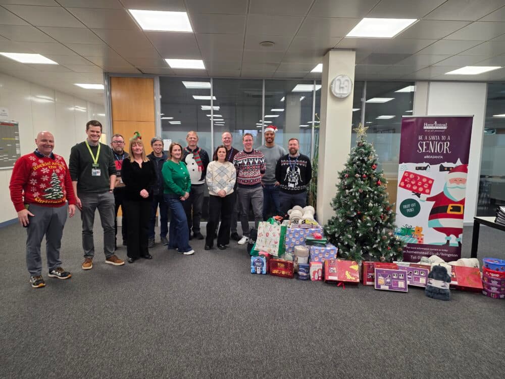 A group of people in festive sweaters stand by a Christmas tree and gifts in an office setting. - Home Instead