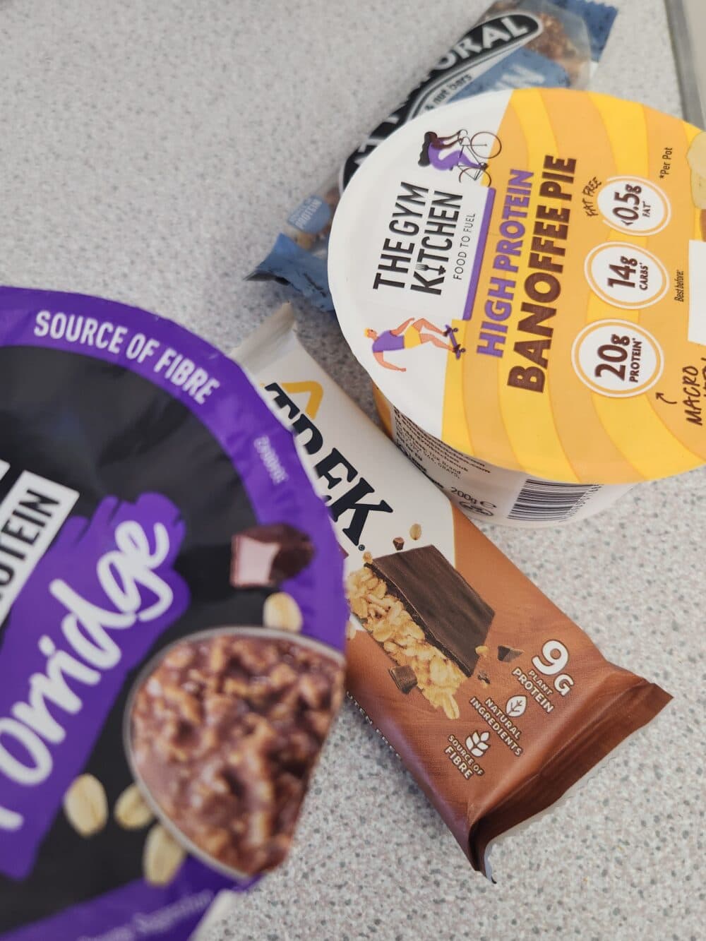 Four high-protein snacks, including bars and porridge, displayed on a light speckled countertop. - Home Instead