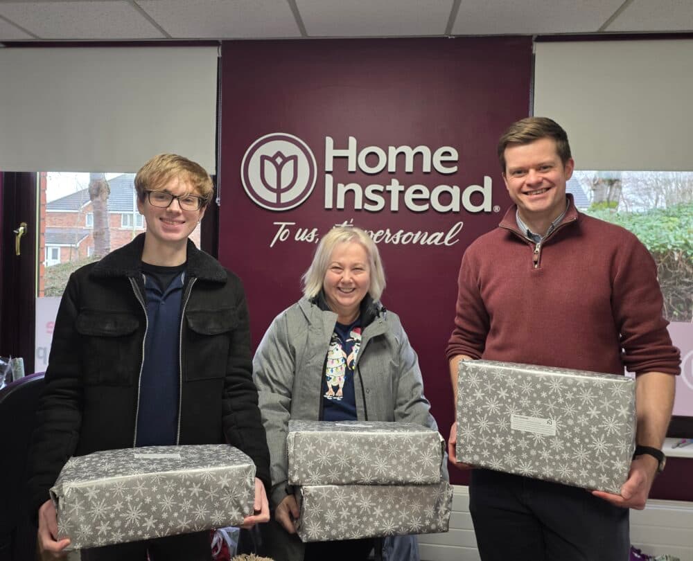Three people smiling and holding gift-wrapped boxes in front of a Home Instead office sign. - Home Instead