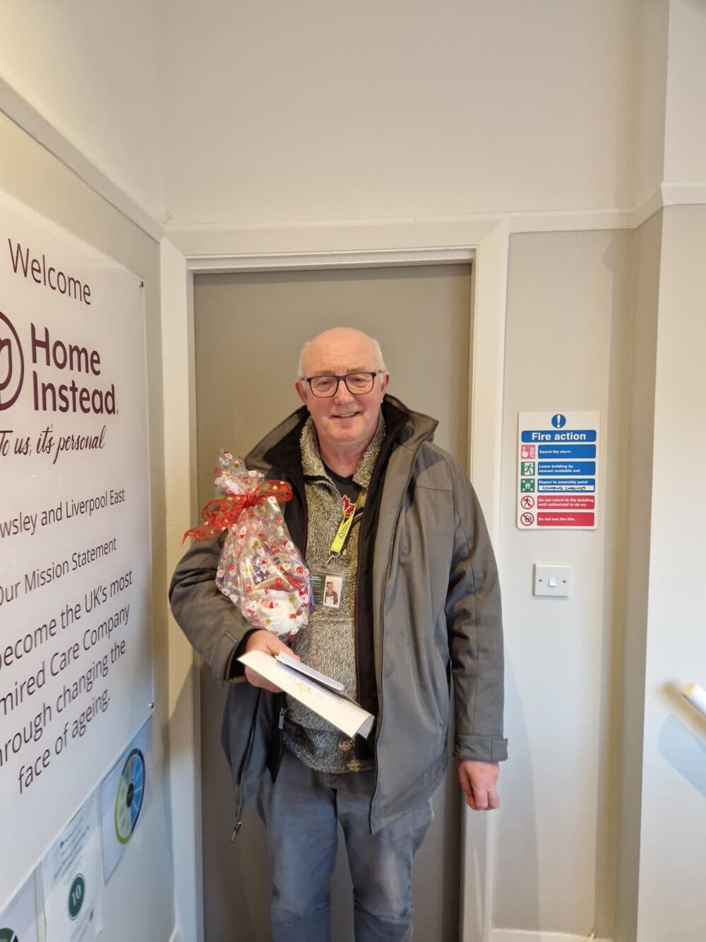 An older man in a coat smiles, holding a wrapped gift and envelope, standing by a "Home Instead" sign. - Home Instead