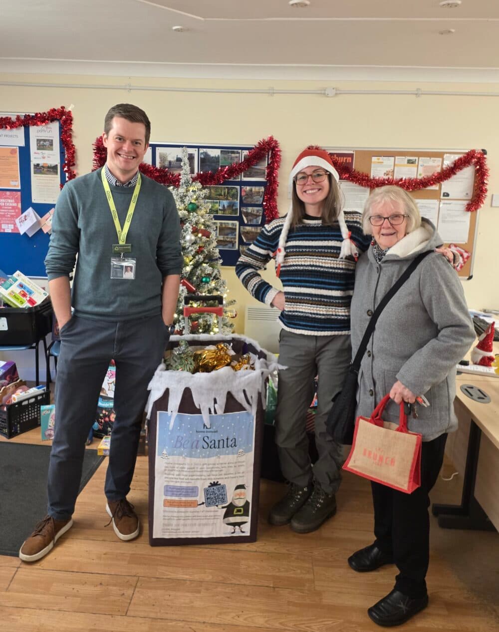 Three people stand smiling by a Christmas tree and donation box in a festive, decorated office. - Home Instead