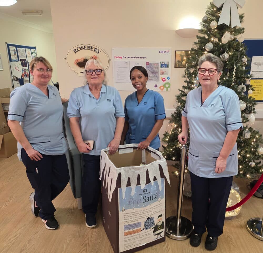 Four women in blue uniforms stand by a Christmas tree and festive donation box, smiling at the camera. - Home Instead