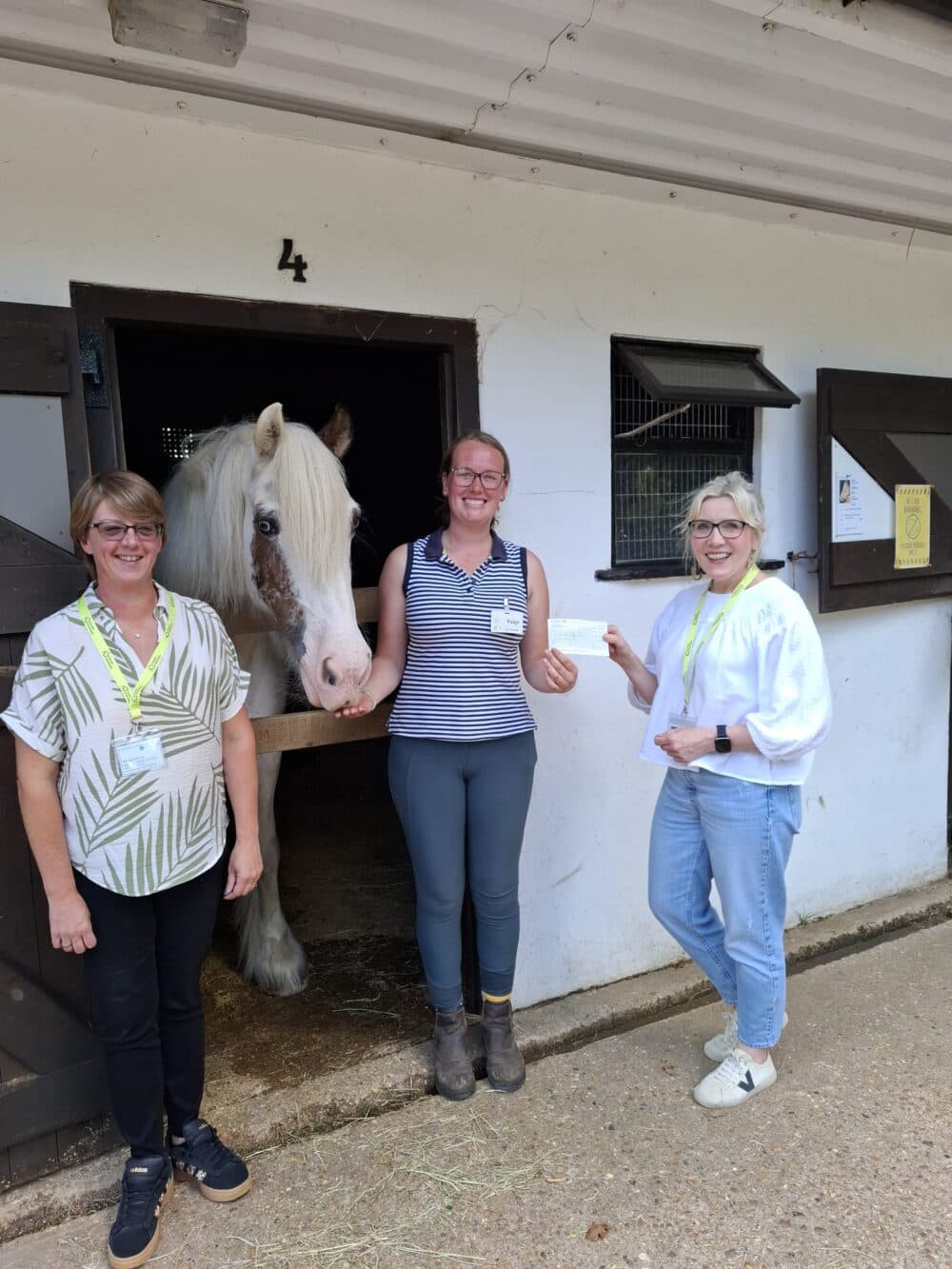 Three women smiling and standing by a white horse in a stable, all wearing name tags. - Home Instead