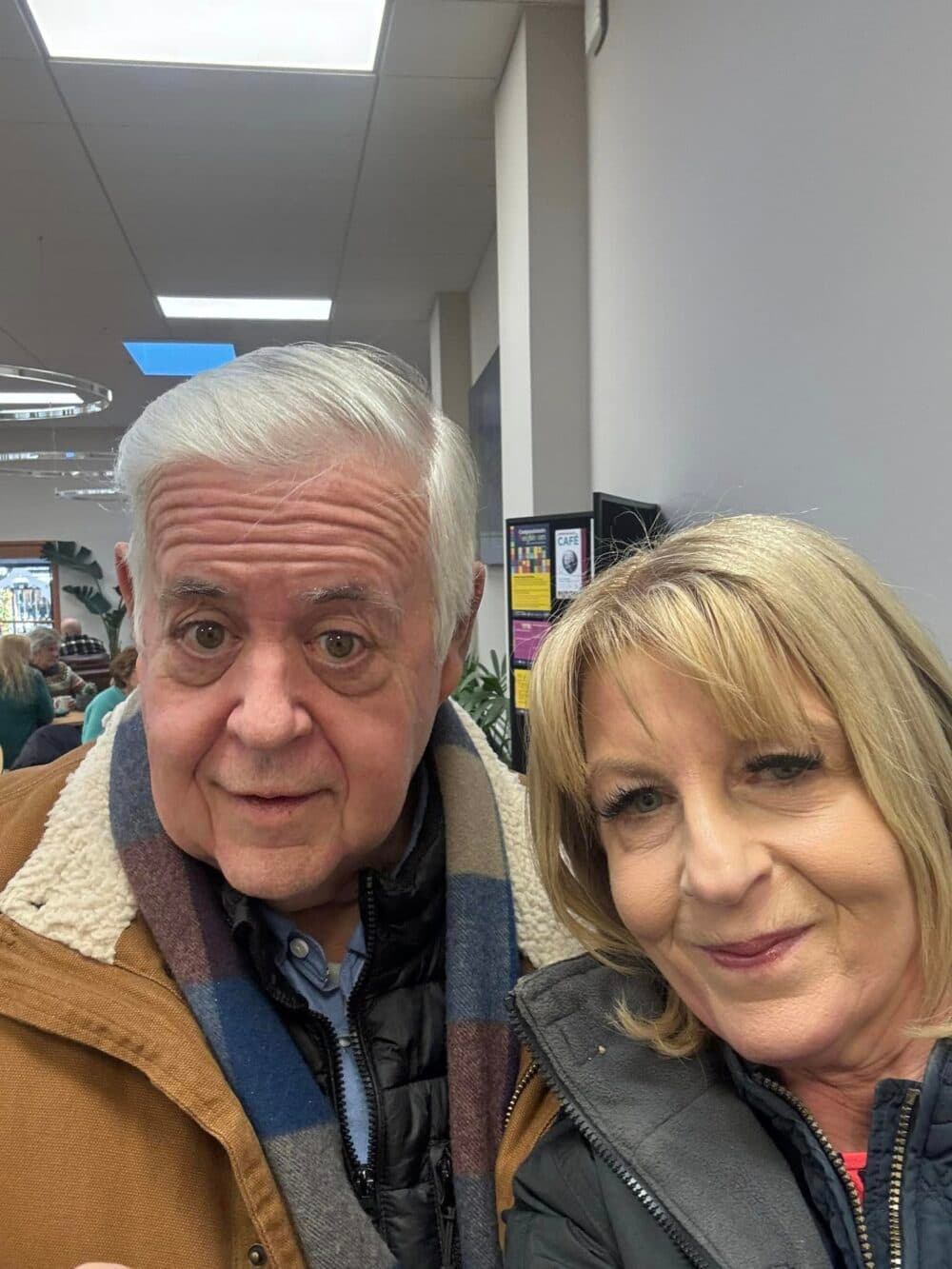An older man and woman smiling for a selfie indoors with people and posters in the background. - Home Instead