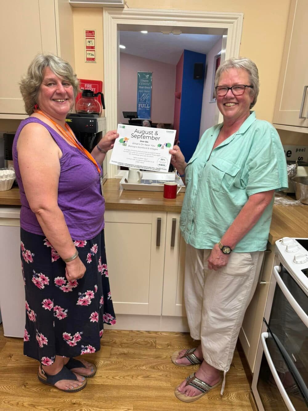 Two smiling women stand in a kitchen, one holding a certificate for "August & September. - Home Instead