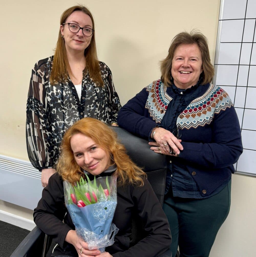 Three women pose indoors; one is seated holding a bouquet of tulips, while the others stand and smile. - Home Instead