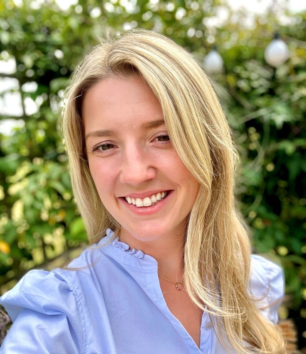 A woman with long blonde hair smiling outdoors, wearing a light blue blouse, with greenery in the background. - Home Instead