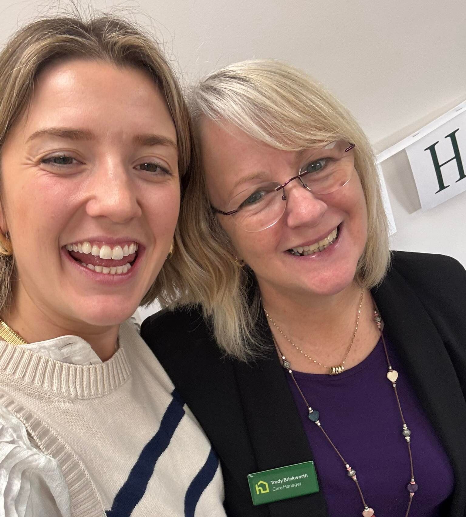Two women smiling together indoors; one wears a badge and glasses, both look happy and friendly. - Home Instead