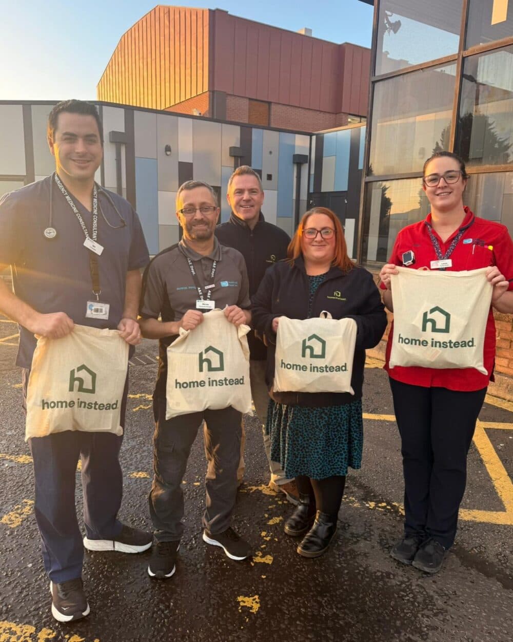 Five smiling people hold "Home Instead" tote bags outside a building on a sunny day. - Home Instead