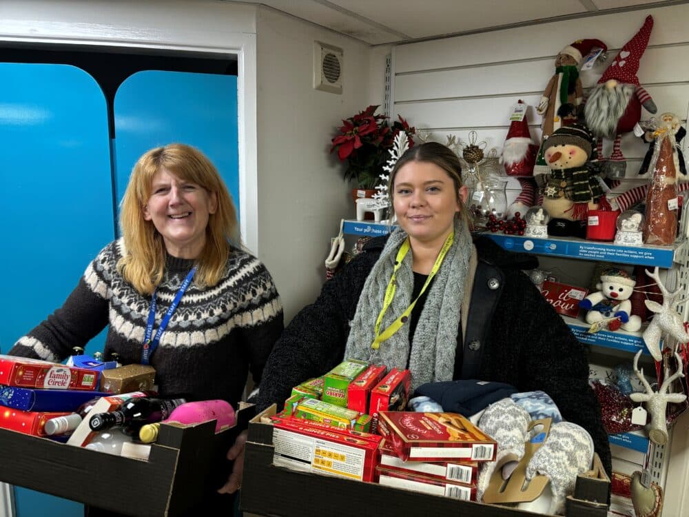 Two women holding boxes of food and gifts, standing in a festive shop decorated with Christmas ornaments. - Home Instead