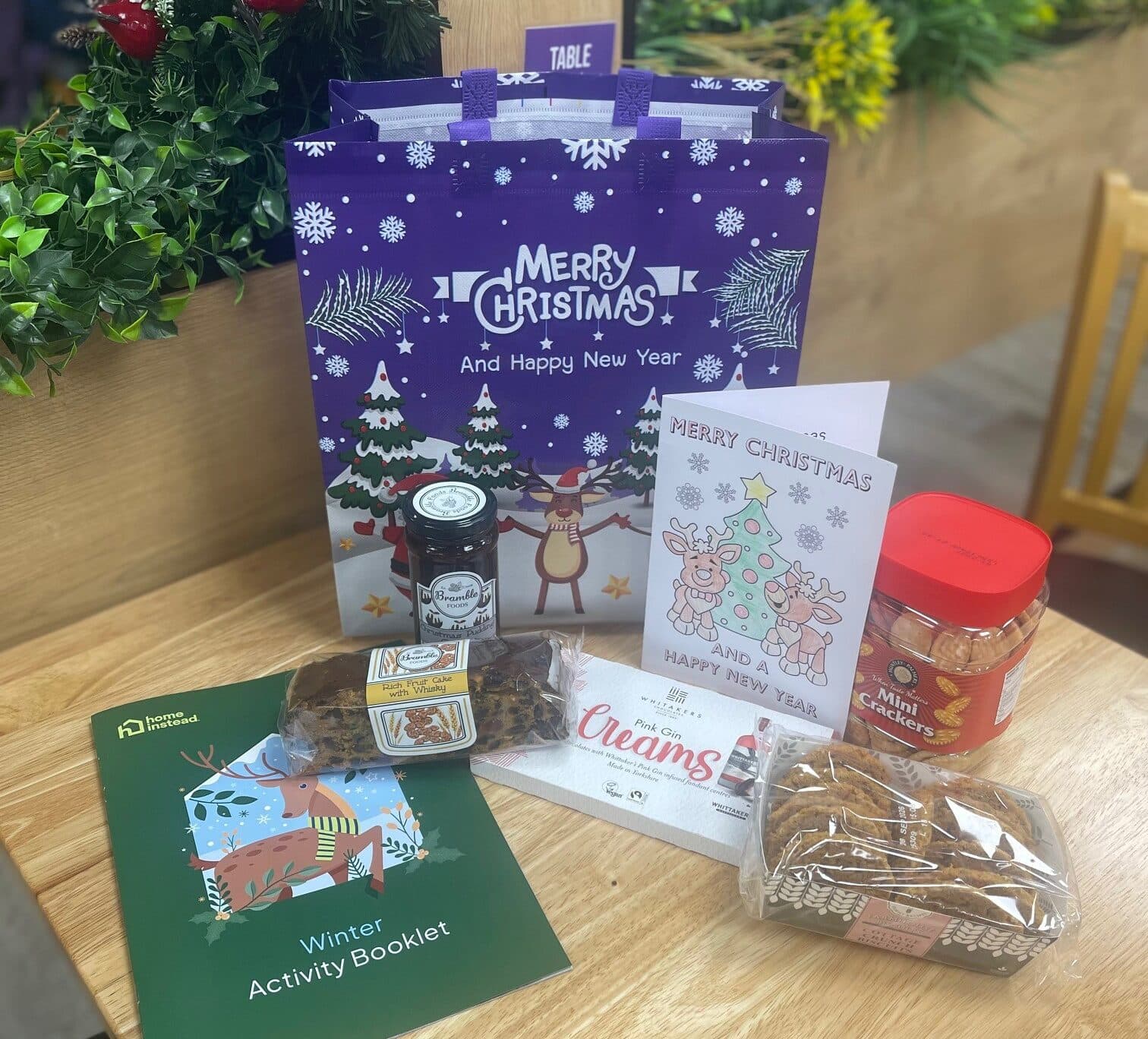 A festive gift bag with holiday treats, a card, and a winter activity booklet on a wooden table. - Home Instead