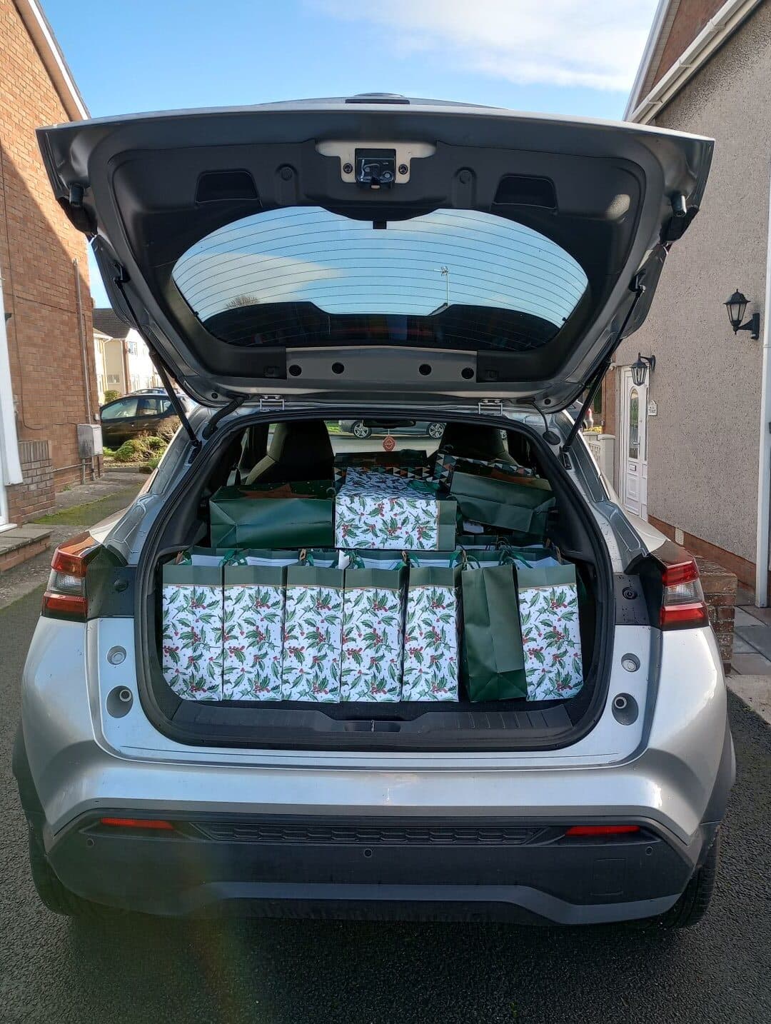 A car trunk filled with festive gift bags decorated with holly patterns, parked in a residential area. - Home Instead