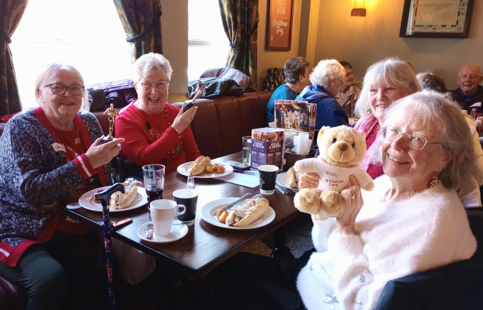 Four smiling elderly women enjoy tea and pastries at a cafe, one holding a teddy bear, another an award. - Home Instead
