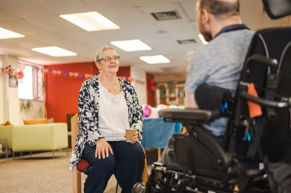 Older woman smiling and talking to a man in a wheelchair in a brightly decorated room. - Home Instead