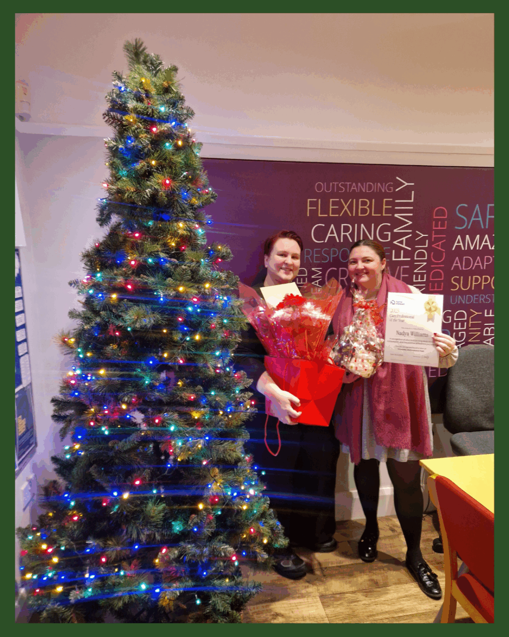 Two people stand by a decorated Christmas tree, holding gifts and a certificate, smiling indoors. - Home Instead