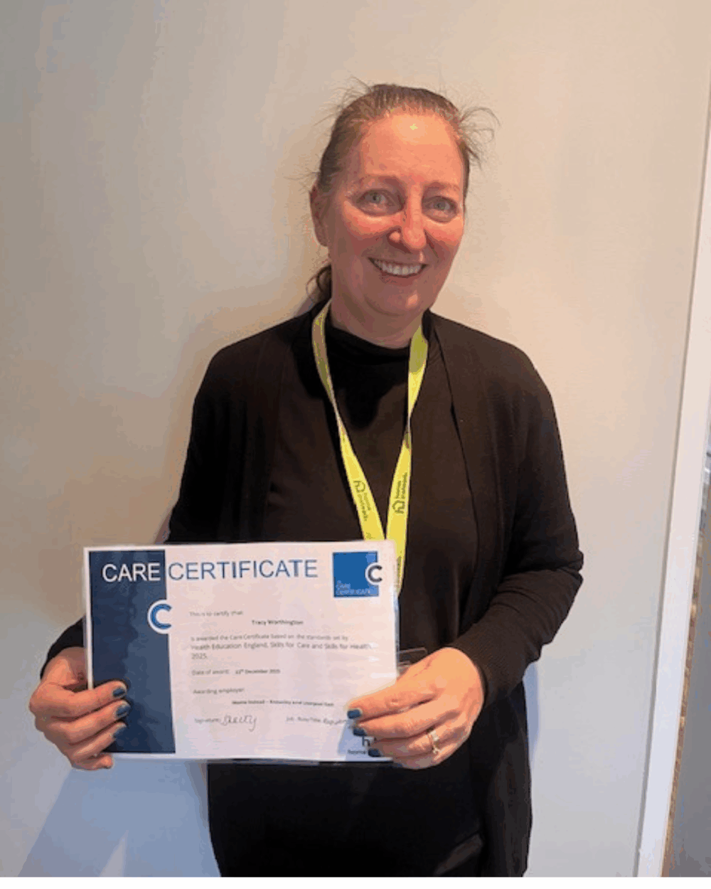Woman smiling and holding a Care Certificate in front of a plain light-colored wall. - Home Instead