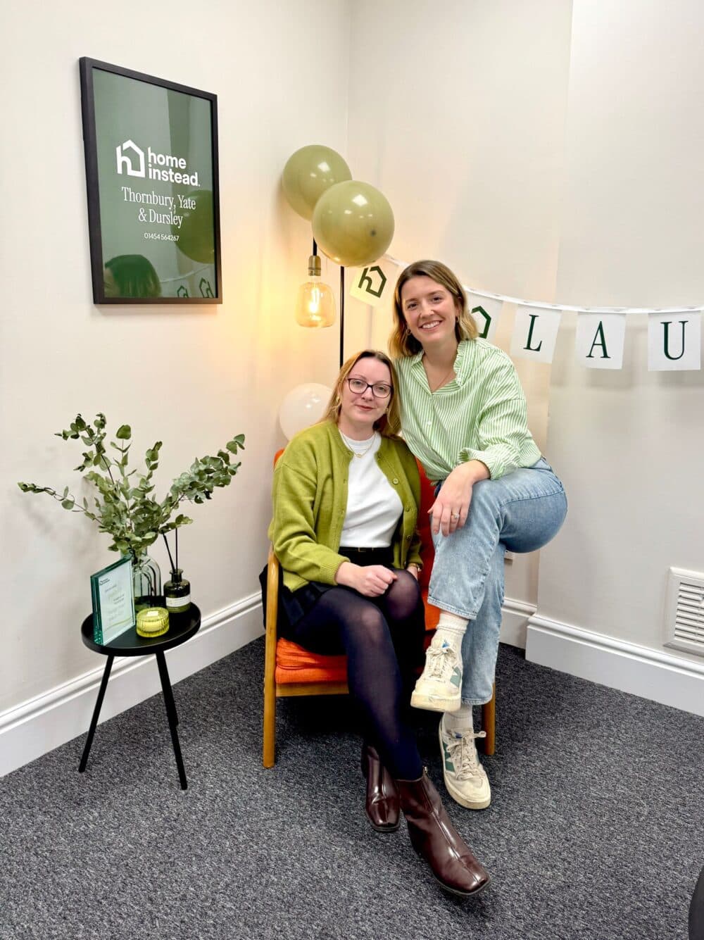 Two women smiling, sitting and posing in a decorated office space with balloons and a "Home Instead" sign. - Home Instead