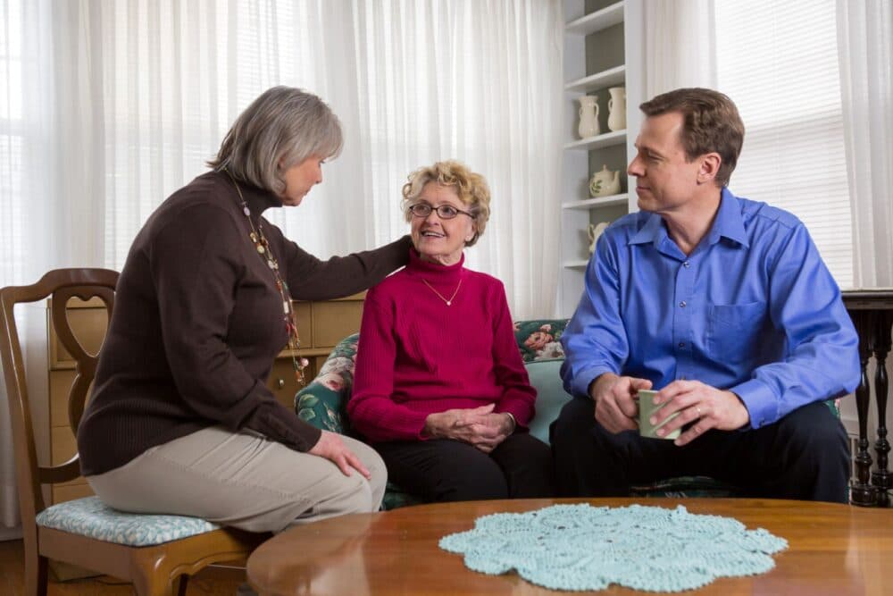 An older woman sits on a couch, talking with a man and a woman in a bright living room. - Home Instead