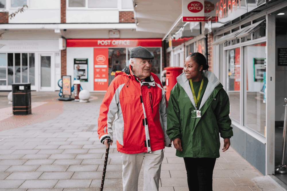 An elderly man with a walking stick walks and talks with a carer on a city street near a post office. - Home Instead