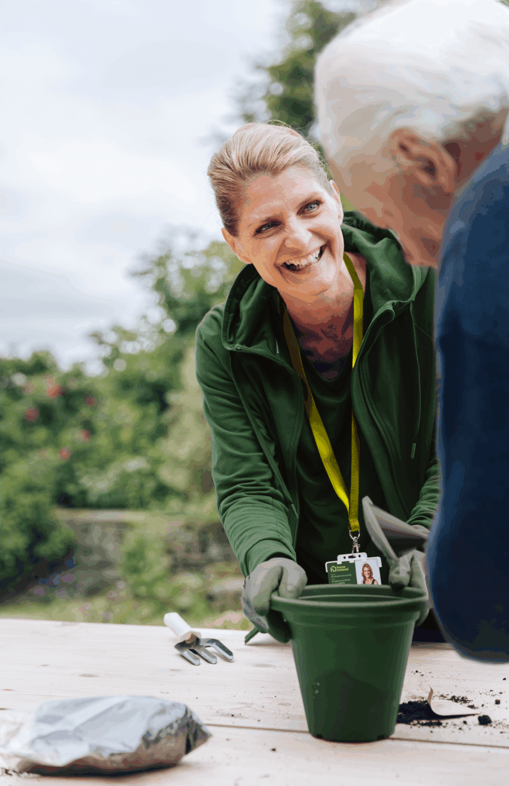 A smiling woman helps an elderly person with gardening, holding a green plant pot outdoors. - Home Instead