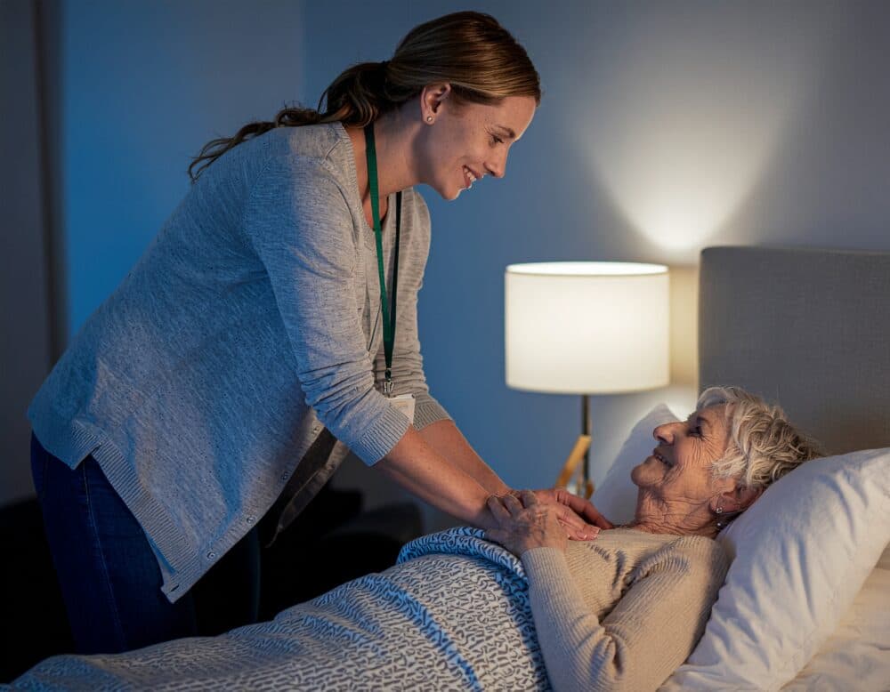A caregiver smiling and comforting an elderly woman who is lying in bed under a blanket. - Home Instead