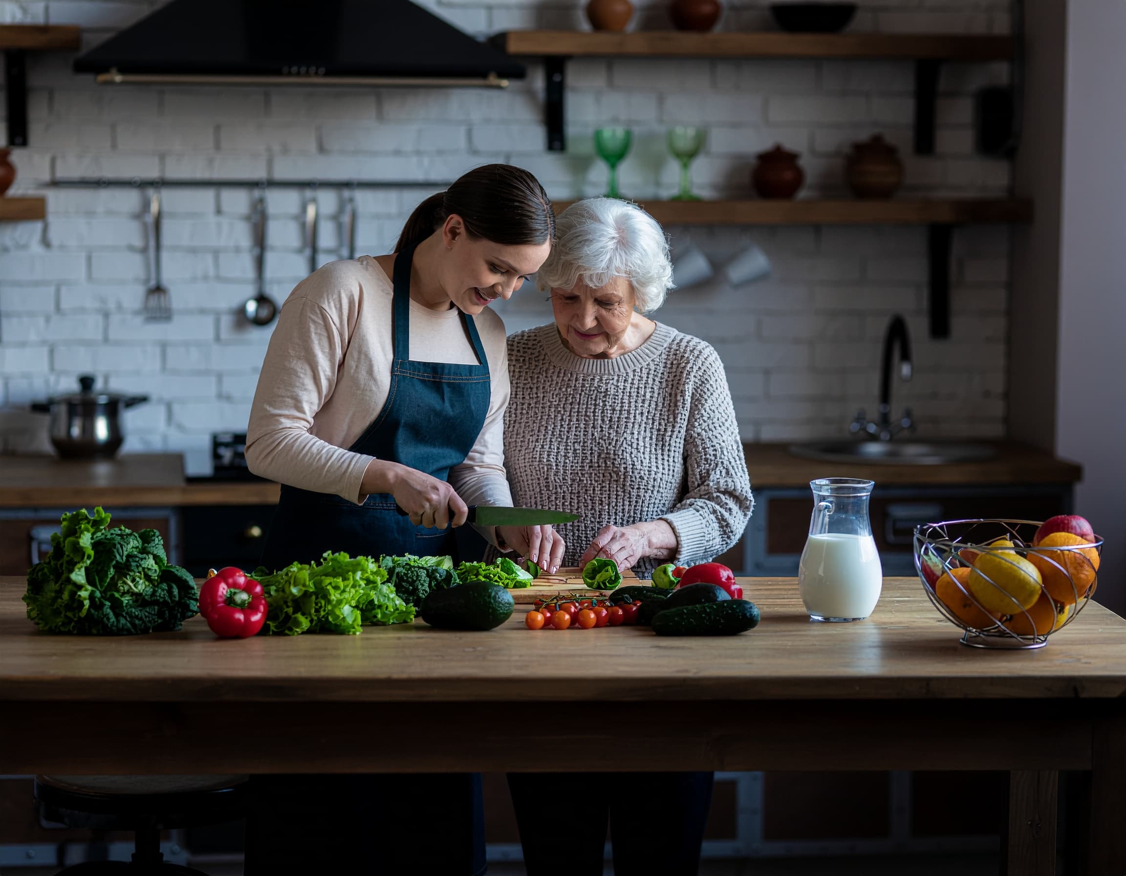 A young woman and an elderly woman chop vegetables together in a kitchen with fresh produce on the worktop. - Home Instead