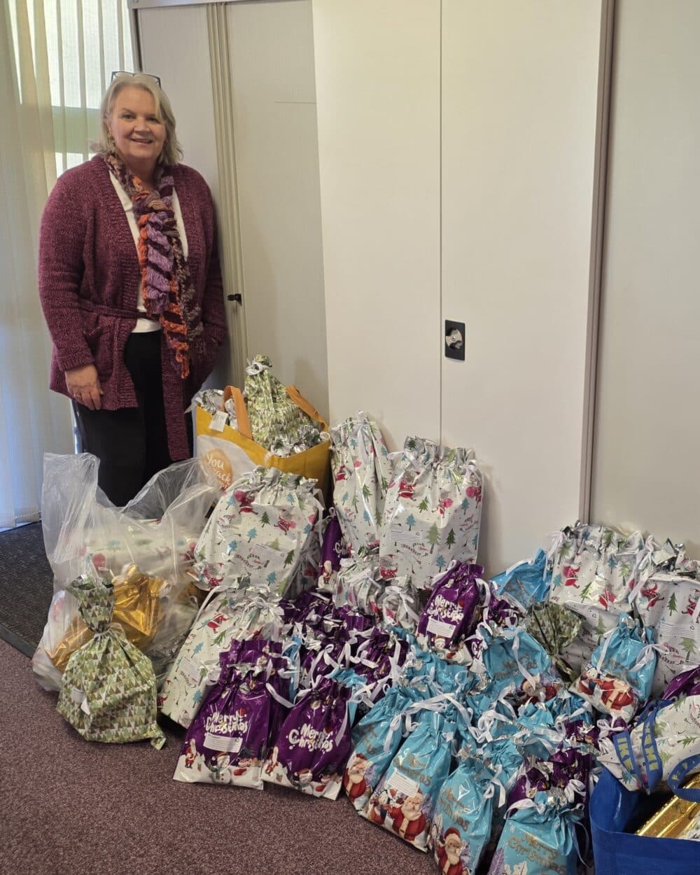 A woman stands next to many colorful gift bags and packages arranged on the floor by a white cabinet. - Home Instead