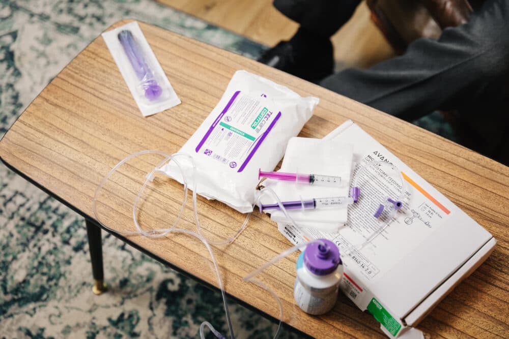 Medical supplies, syringes, tubing, and medication on a wooden table, with part of a person's legs visible. - Home Instead