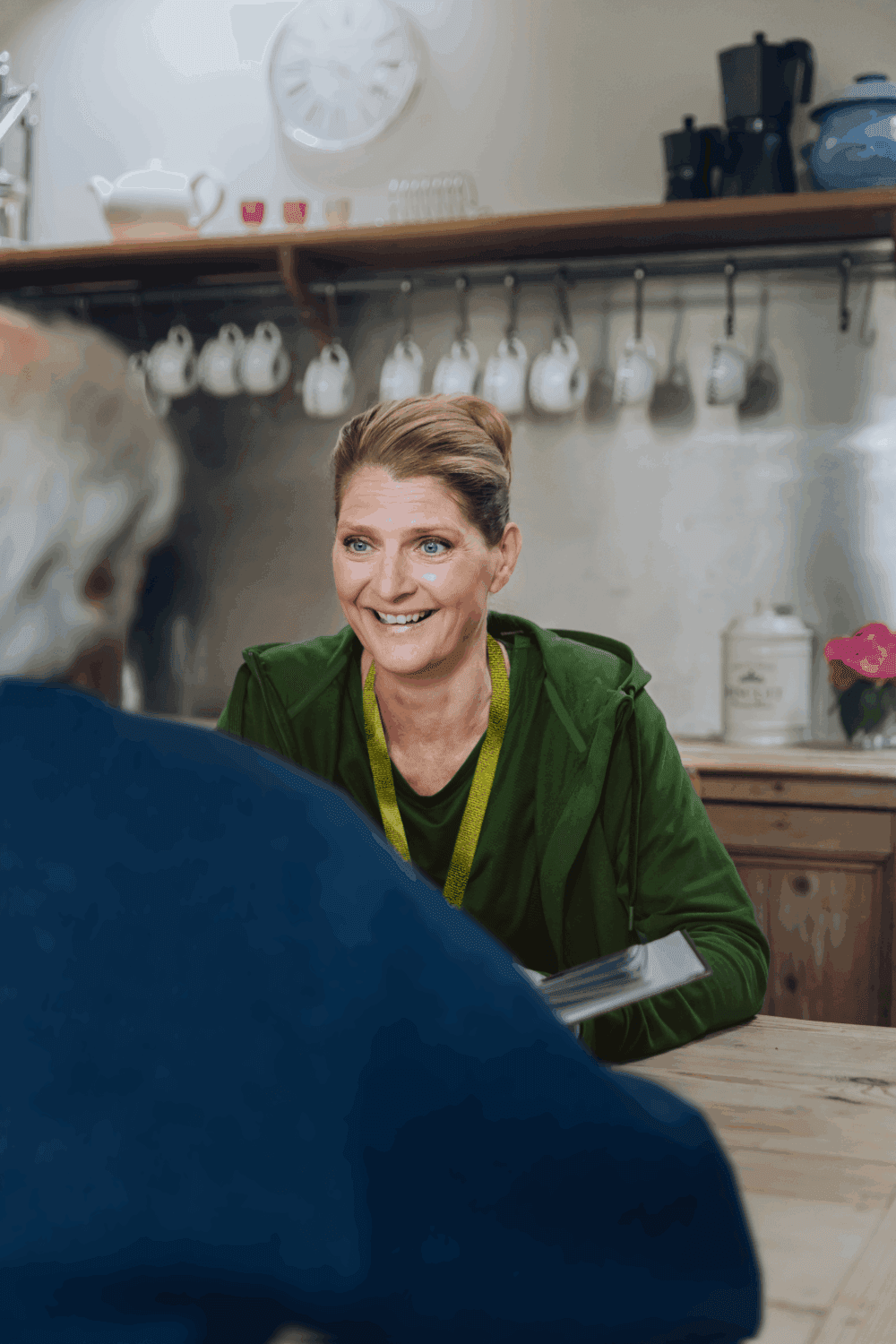 A smiling woman sits at a kitchen table, talking to an older man, with mugs and teapots in the background. - Home Instead