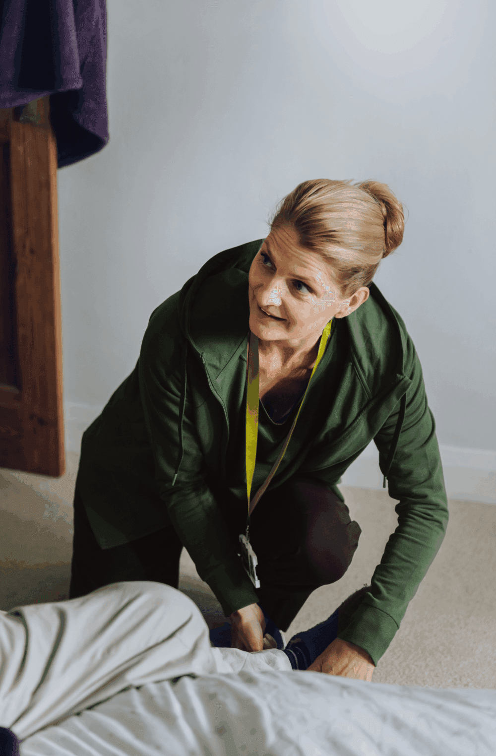 A woman in a green jacket helps someone put on shoes next to a bed, smiling warmly. - Home Instead