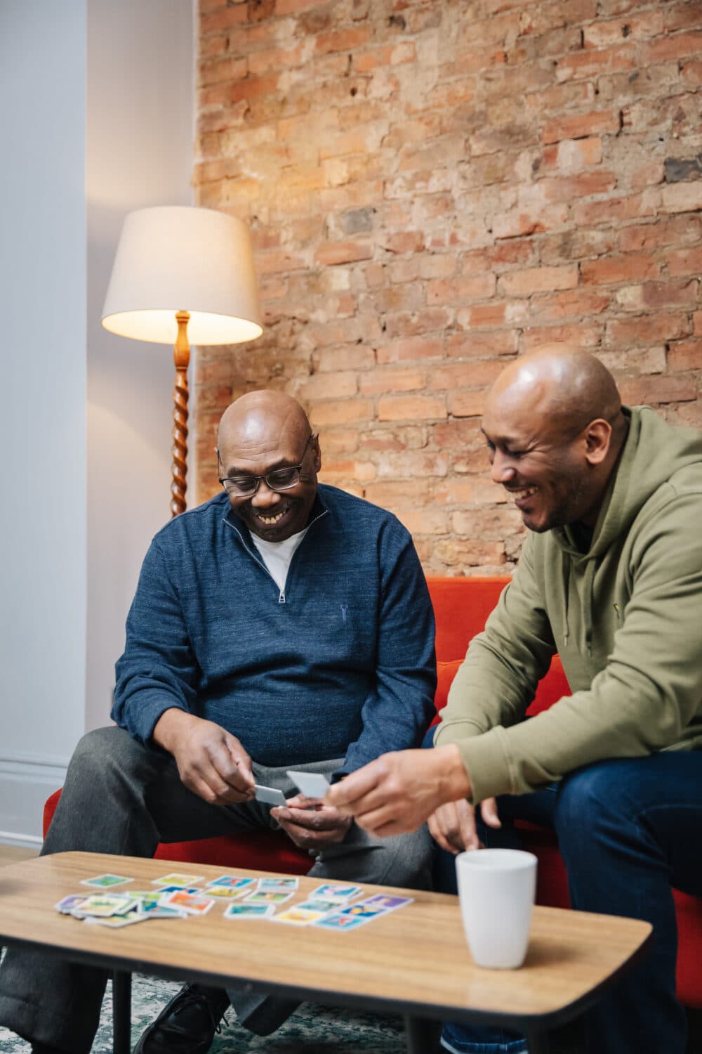 Two men sitting on a couch, smiling and playing a card game on a coffee table in a cozy living room. - Home Instead
