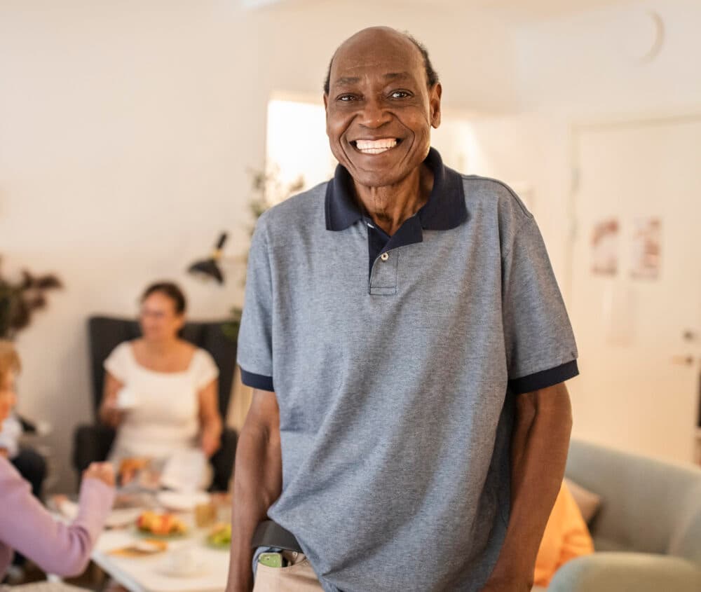 Smiling older man stands indoors; people sit and talk in the blurred background. - Home Instead
