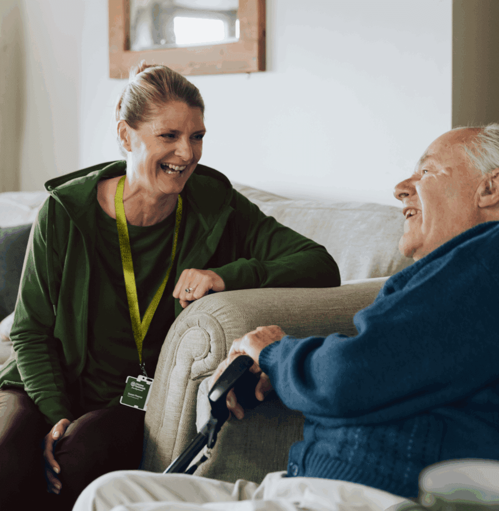 A carer smiles and chats with an elderly man sitting on a sofa, both appearing happy and relaxed. - Home Instead