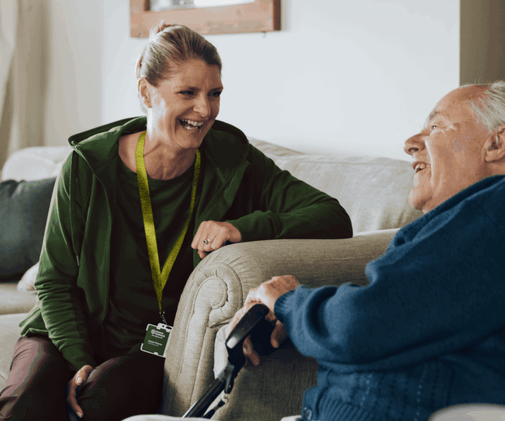 A smiling carer talks with an elderly man sitting on a sofa, holding a walking stick. - Home Instead