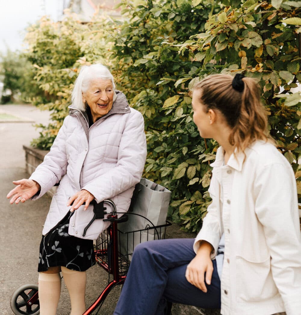 Elderly woman with a walker and a younger woman chat outside by green bushes. - Home Instead