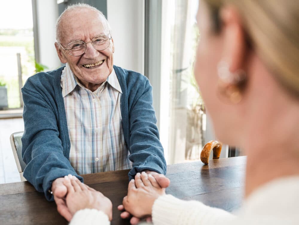 Smiling elderly man holding hands across a table with a woman in a bright, sunlit room. - Home Instead