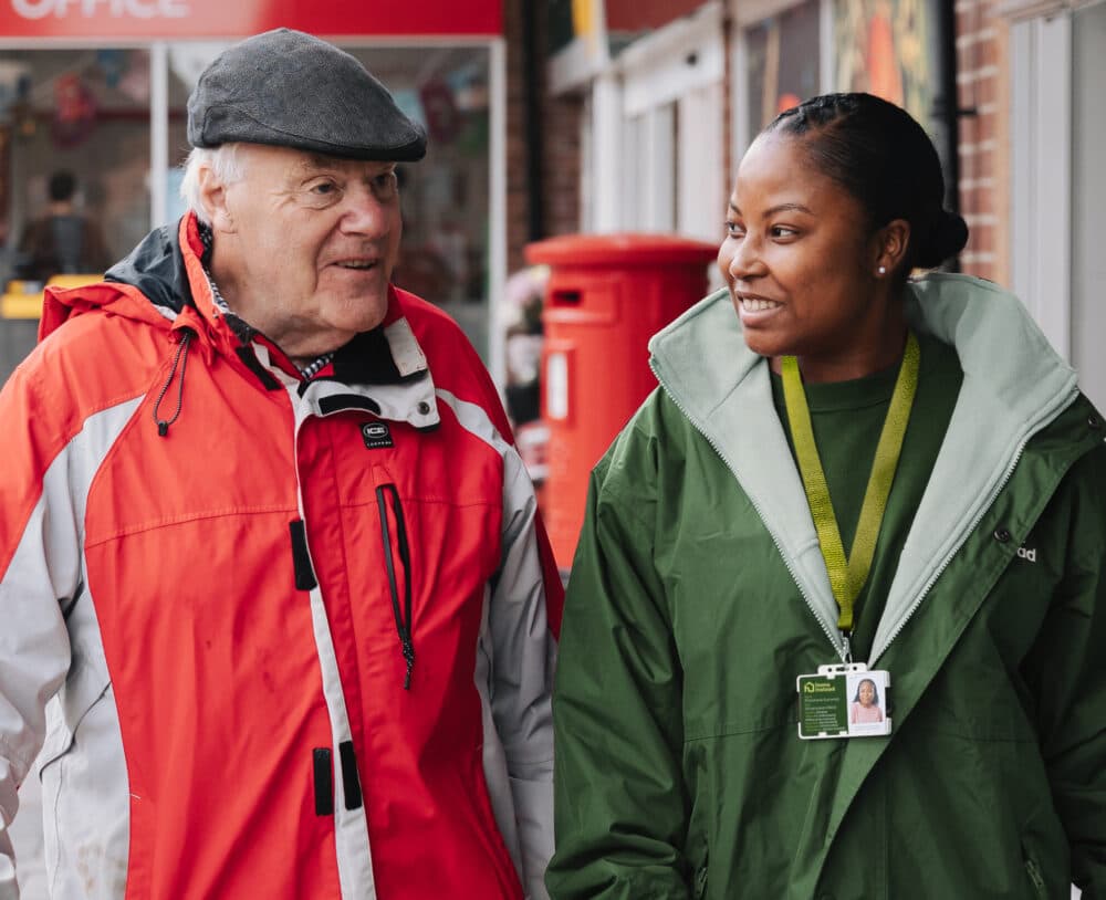 An elderly man and a woman in uniforms smiling and talking outside near a red pillar box. - Home Instead