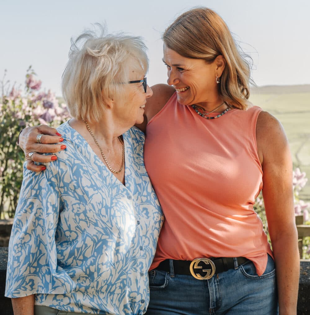 Two women smiling at each other outdoors, with arms around each other, flowers and fields in the background. - Home Instead