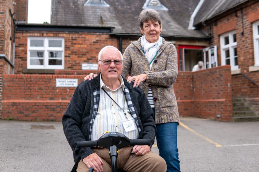 An older man on a mobility scooter and a woman stand outside a red-brick building, smiling at the camera. - Home Instead